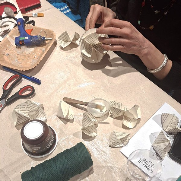 Person assembling a spherical ornament from folded paper segments with printed text; crafting workspace includes a hot glue gun, scissors, pen, spool of green string, small container with glue and brush, and a disposable coffee cup; several folded paper pieces are laid out as the person carefully positions them