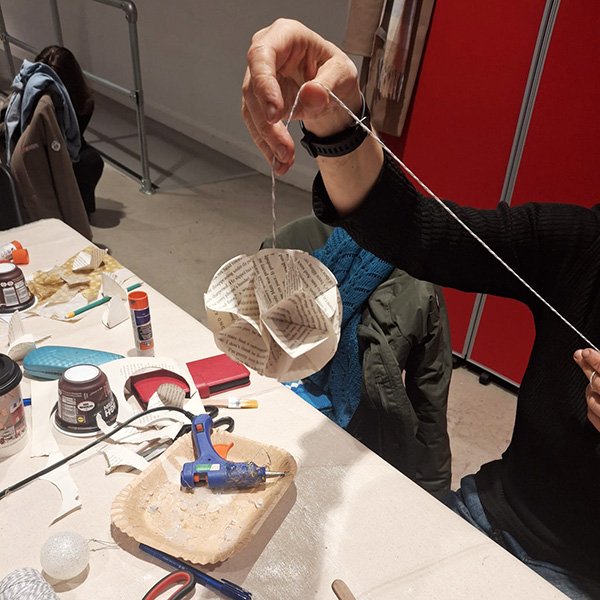 Person holding a spherical hanging ornament made from folded paper segments with printed text; tabletop includes glue sticks, hot glue gun, scissors, paper pieces, and containers of adhesive, suggesting a hands-on craft activity using recycled materials