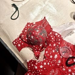 Festive crafting scene with a red glitter-covered Christmas ornament partially wrapped in red tissue paper printed with white stars and 'Merry Christmas' text; surrounding materials include a container of red glitter, green string or ribbon, and white tissue paper on a light-colored fabric surface
