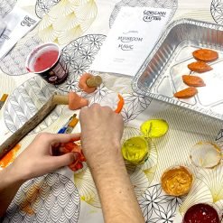 Hands shaping orange clay mushrooms during a “Mushroom Magic” crafting workshop, surrounded by colorful paints, tools, and a journal titled “Conscious Crafting.