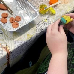 Hands painting a polymer clay mushroom cap with bright yellow and teal acrylic paint using a sponge applicator, next to a foil tray holding unfinished orange clay caps with holes, on a patterned tablecloth with open paint pots
