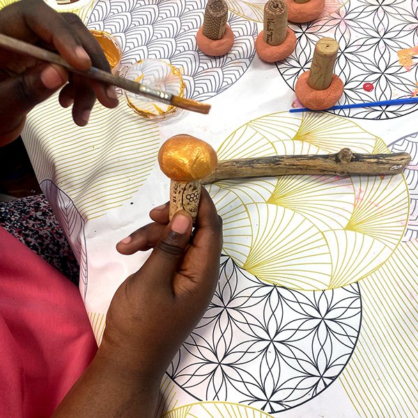 Hands painting a metallic gold finish onto a polymer clay mushroom cap attached to a cork stem, using a paintbrush dipped in gold paint, on a patterned tablecloth with other unfinished clay mushrooms in the background