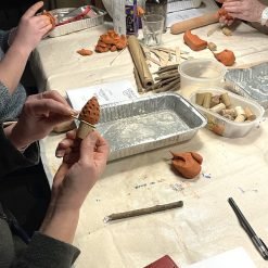 Hands assembling a handmade polymer clay mushroom ornament by attaching a textured orange clay cap to a cork stem, on a busy crafting table with twigs, clay pieces, tools, wine corks, and other participants working in the background