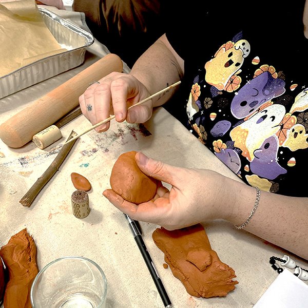 Hands shaping orange polymer clay into a mushroom cap while inserting a twig stem, on a cluttered crafting table with a rolling pin, clay pieces, tools, and a glass of water