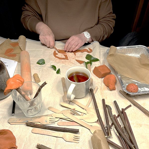 "Person crafting handmade mushroom decorations from polymer clay at a cozy workspace table, surrounded by orange clay pieces, twigs for stems, wooden tools, green leaves, a rolling pin, and a mug of tea"