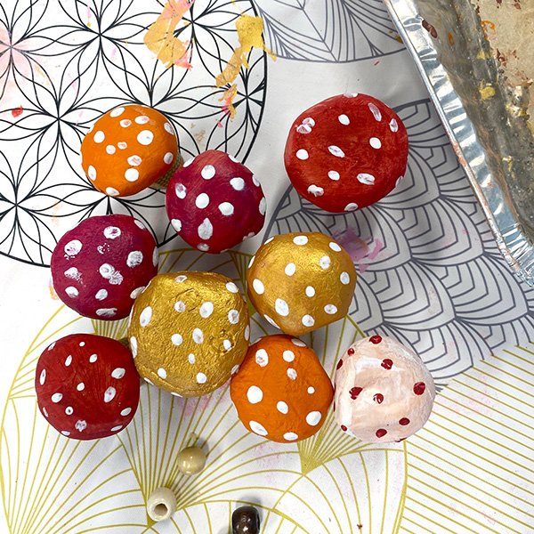 "Hand-painted mushroom cap beads in vibrant autumn colors—red, orange, purple, and metallic gold—with white polka dots, arranged on a patterned workspace with paint splatters and crafting materials"