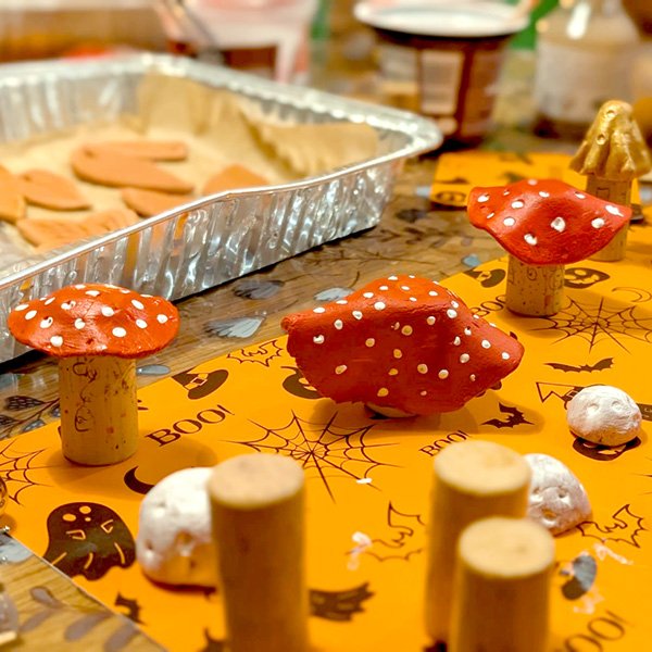 Handmade clay mushrooms with red caps and white dots, crafted on an orange Halloween-themed mat featuring spiders, bats, and “BOO!”—with unfinished pieces and a tray of cutouts in the background.