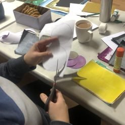 Person cutting circular shapes from white paper at craft table with colored paper, glue stick, and snacks.