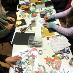 Group crafting collage-style art with colored paper, glue sticks, and nature-themed cut-out shapes at table.