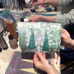 Person holding floral monoprint in green, white, and purple at art table with paint supplies.