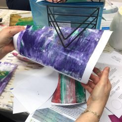 Person holding abstract artwork in purple, green, and white at table with art supplies and printed sheets.