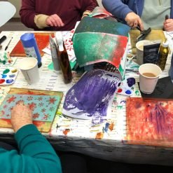 Group creating colorful prints with rollers, stencils, and paint at table covered in art supplies.